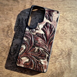 May include: A rectangular smartphone case featuring a black frame and a textured surface with a deep brown and cream-colored embossed floral pattern. The design consists of intricate, swirling leaf-like shapes that cover the back of the case.