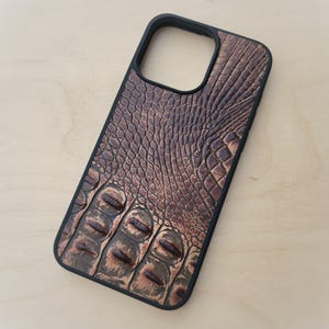 May include: A rectangular smartphone case featuring a black frame and a textured back panel with a brown, embossed crocodile skin pattern. The design includes a large cutout for a camera lens array at the top and a detailed, raised scale texture.