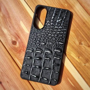 May include: A black smartphone case featuring a textured crocodile skin pattern. The design includes a prominent, raised scale texture across the entire back surface and a large, oval-shaped cutout at the top for the camera lens assembly.