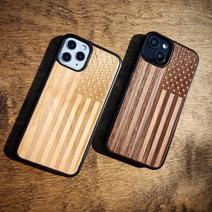Könnte beinhalten: Zwei Smartphone-Hüllen mit schwarzem Rahmen und hölzernen Rückseiten, die mit einem eingravierten amerikanischen Flaggendesign versehen sind. Die linke Hülle besteht aus hellem Holz, die rechte aus dunklerem Holz, beide mit Kameraaussparungen.