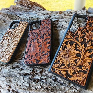 May include: Three rectangular phone cases with black edges and wood-toned back panels featuring intricate, deeply carved floral and vine patterns. The cases vary in size and wood colour, ranging from a light natural finish to a dark, rich brown stain.