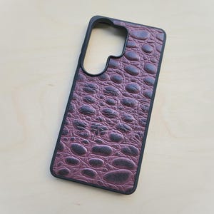 May include: A rectangular smartphone case featuring a dark purple, textured faux-alligator skin pattern on the back panel. The case has a solid black frame and a large, irregular cutout at the top left corner designed to accommodate a camera module.