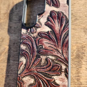 May include: A phone case with a detailed floral design in shades of brown and burgundy. The case has a black frame and a textured, embossed pattern. The design is reminiscent of vintage leatherwork.