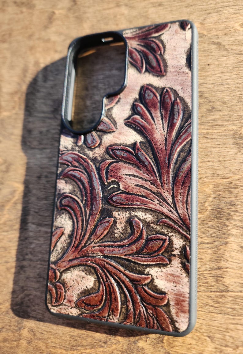 May include: A phone case with a detailed floral design in shades of brown and burgundy. The case has a black frame and a textured, embossed pattern. The design is reminiscent of vintage leatherwork.