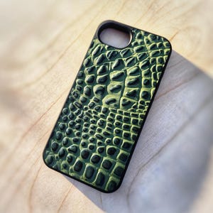May include: A rectangular phone case featuring a textured, raised green and black crocodile skin pattern. The case has a black border and a circular cutout at the top for a camera lens. The surface of the case displays a detailed, repeating scale design.