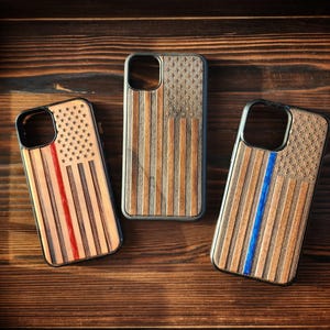 May include: Three rectangular phone cases featuring wood grain textures and engraved American flag designs. One case includes a vertical red stripe, one features a standard wood-toned flag, and one displays a vertical blue stripe on the flag pattern.