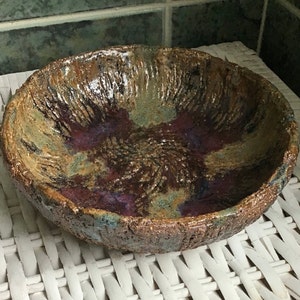 May include: A round ceramic bowl with a textured, uneven rim and a rough, cratered exterior surface. The interior features a mottled glaze in shades of deep purple, earthy brown, and muted green, with a dark, glossy finish in the centre.