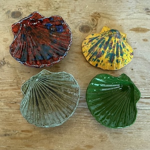 May include: Four ceramic dishes shaped like scallop shells. One is glossy red with dark blue accents, one is yellow with blue speckles, one is a muted sage green, and one is a solid, vibrant emerald green. Each shell features radial ridges.