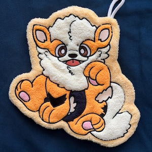 May include: A plush, textured wall hanging shaped like a stylized orange and white creature with large brown eyes, a small black nose, and a pink tongue. The design features black accents on the body, pink paw pads, and a white hanging loop.