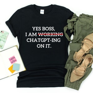 Op de afbeelding: Zwart T-shirt met witte tekst die "YES BOSS, I AM WORKING CHATGPT-ING ON IT." luidt. Het woord "WORKING" is doorgestreept met een rode lijn.