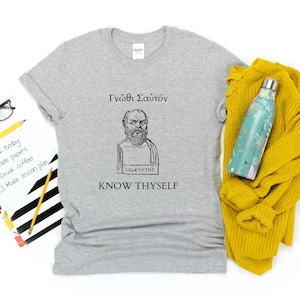 Può includere: Una maglietta grigio chiaro con un'illustrazione in bianco e nero di Socrate e la frase greca «Conosci te stesso». Un maglione giallo e una borraccia turchese sono accanto alla maglietta. La maglietta è di cotone.