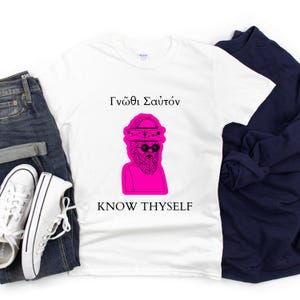 Può includere: T-shirt bianca con grafica rosa di un busto con occhiali da sole e cappello. La frase greca "Γνῶθι Σαυτόν" è sopra la grafica e la frase inglese "KNOW THYSELF" sotto. La maglietta è abbinata a jeans blu e scarpe da ginnastica bianche.