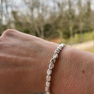 Könnte beinhalten: Ein silbernes Armband aus kleinen, strukturierten ovalen Perlen.