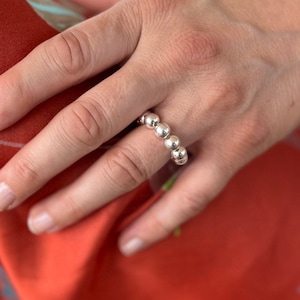 Könnte beinhalten: Ein silberner Ring mit fünf kugelförmigen Perlen. Der Ring wird an einem Finger getragen, vor einem roten Stoffhintergrund. Das Design des Rings ist schlicht und elegant.