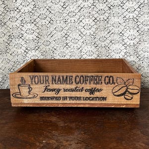 May include: A rectangular wooden box with the text "YOUR NAME COFFEE CO." and "Fancy roasted coffee BREWED IN YOUR LOCATION" etched on the front. The box features a coffee cup, saucer, and coffee bean design.