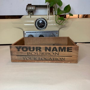 May include: A wooden box with the text "YOUR NAME BOURBON YOUR LOCATION" printed on the front. The box is brown and has a rustic look.