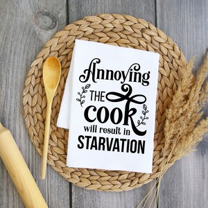 May include: White kitchen towel with black text that reads "Annoying the cook will result in starvation". The towel is on a woven straw placemat with a wooden spoon and rolling pin beside it.
