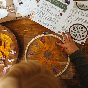 May include: A craft project showing a dried yellow flower pressed in an embroidery hoop. Yellow petals and purple flowers are scattered around the hoop on a wooden surface. Instructions and mushroom illustrations are visible.