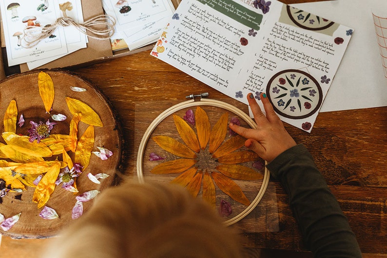 May include: A craft project showing a dried yellow flower pressed in an embroidery hoop. Yellow petals and purple flowers are scattered around the hoop on a wooden surface. Instructions and mushroom illustrations are visible.