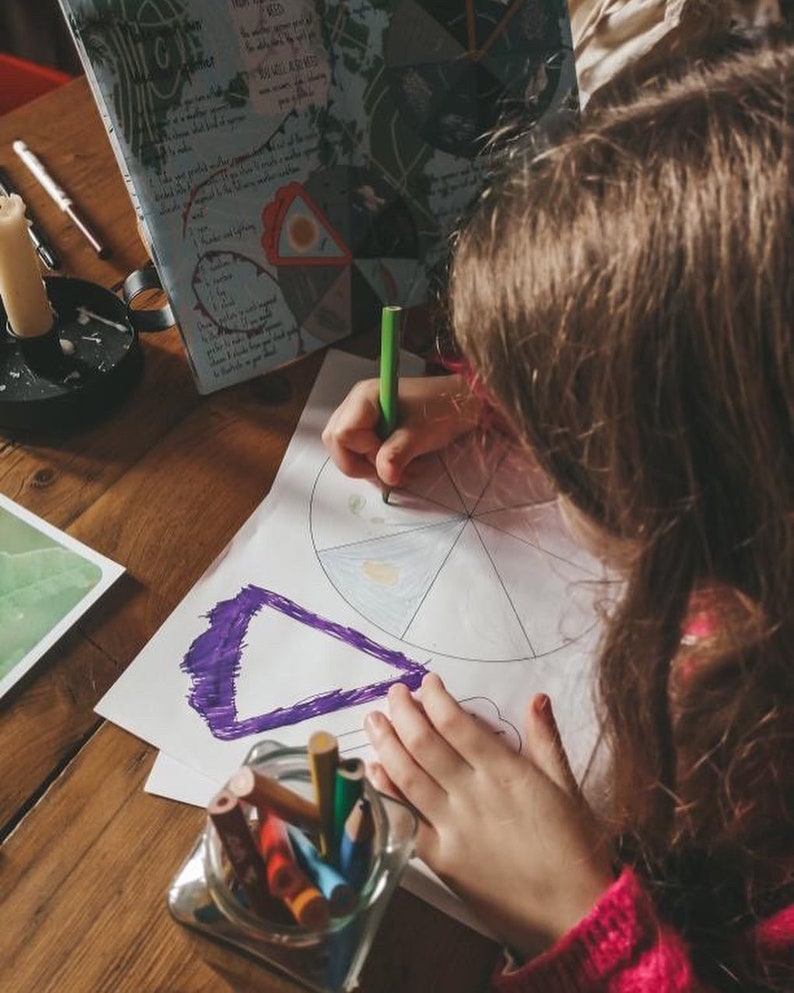 Puede incluir: Un ni&ntilde;o dibuja en papel blanco con un l&aacute;piz verde, coloreando un diagrama circular. Un tarro de cristal con l&aacute;pices de colores est&aacute; sobre una mesa de madera. Una vela en un portavelas negro y un libro con texto manuscrito tambi&eacute;n est&aacute;n sobre la mesa.