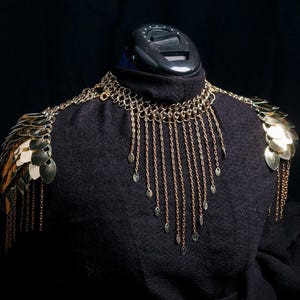 May include: Gold-toned shoulder armour with a chainmail collar and hanging chains. The armour has scale-like plates on the shoulders and dangling teardrop charms. It's displayed on a black mannequin head against a dark background.