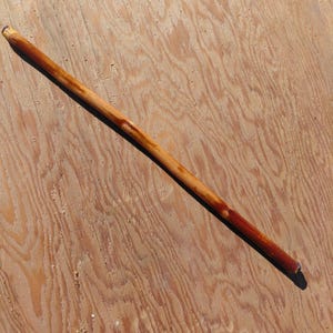 May include: A long, wooden walking stick with a natural finish. The stick is primarily light brown with darker reddish-brown accents near the ends. The wood grain is visible, and the stick appears sturdy and well-crafted.