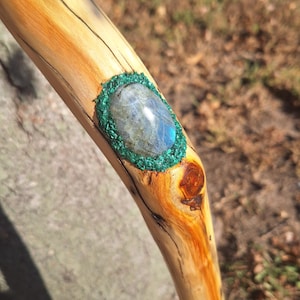 May include: A wooden walking stick with a light brown finish. A large oval-shaped gemstone is set into the stick, surrounded by a green border. The gemstone is a light blue color.
