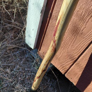 K&ouml;nnte beinhalten: Ein h&ouml;lzerner Wanderstock mit nat&uuml;rlicher Oberfl&auml;che, gebogenem Design und schwarzer Gummispitze. Das Holz hat eine hellbraune Farbe mit roten Streifen und dunklen Maserungsmustern. Der Stock lehnt an einer verwitterten roten und wei&szlig;en Holzkonstruktion.