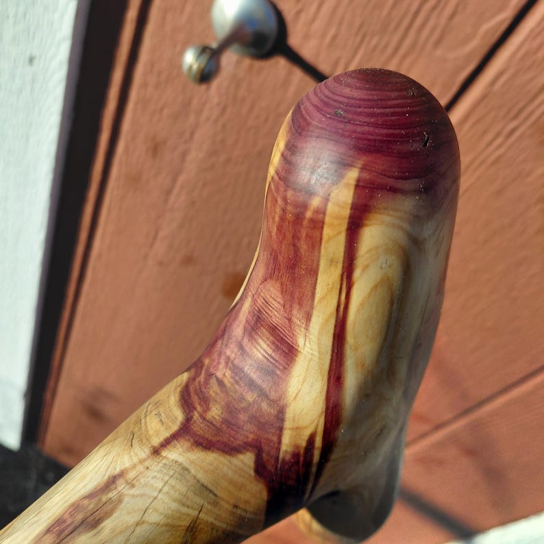 K&ouml;nnte beinhalten: Nahaufnahme eines handgefertigten Holzobjekts mit geschwungener Form. Das Holz weist ein nat&uuml;rliches Maserungsmuster mit einer Mischung aus hellgelben und tiefvioletten Farbt&ouml;nen auf. Das Objekt scheint Teil eines gr&ouml;&szlig;eren St&uuml;cks zu sein.