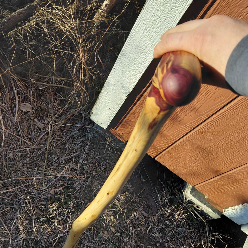 K&ouml;nnte beinhalten: Handgefertigter Wanderstock aus Holz mit einem abgerundeten, rot-violett und gelb get&ouml;nten Griff. Der Schaft ist hellbraun. Der Wanderstock lehnt an einer braunen und wei&szlig;en Holzkonstruktion.