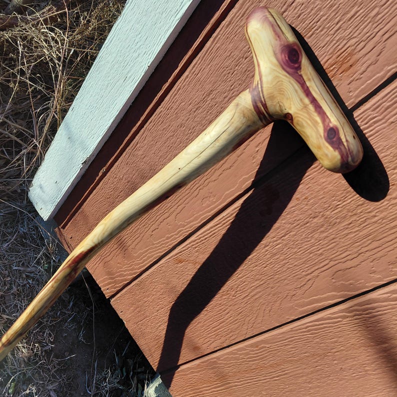 K&ouml;nnte beinhalten: Ein handgefertigter Holzspazierstock mit einem T-f&ouml;rmigen Griff. Der Griff weist ein auff&auml;lliges Muster aus hellem Holz mit tiefvioletten Akzenten auf. Der Schaft ist in einem helleren Holzton gehalten. Der Stock wirkt robust und gut verarbeitet. Ca. 100 cm lang.