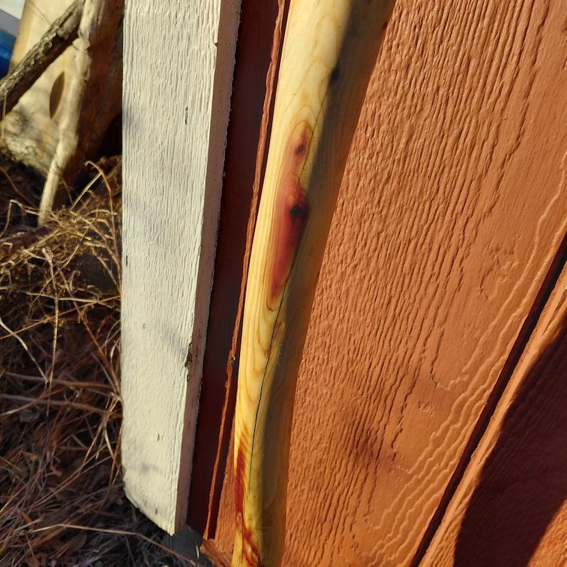 K&ouml;nnte beinhalten: Axtstiel aus hellem Holz mit einem rotbraunen Abschnitt. Der Griff ist leicht gebogen und vor einer strukturierten, rotbraunen Holzoberfl&auml;che und einer wei&szlig; lackierten Holzoberfl&auml;che platziert.