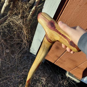K&ouml;nnte beinhalten: Handgeschnitzter Gehstock aus Holz mit gebogenem Griff. Der Stock weist ein nat&uuml;rliches Holzmaserungsmuster mit r&ouml;tlich-braunen Akzenten und Knoten auf. Der Griff ist ergonomisch geformt.