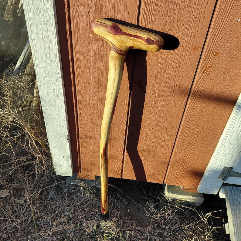 K&ouml;nnte beinhalten: Ein handgefertigter Holzspazierstock mit gebogenem Griff. Der Griff ist hellbraun mit rot-violetten Akzenten. Der Schaft ist ein hellerer Braunton. Der Stock lehnt an einer braunen Holzwand.