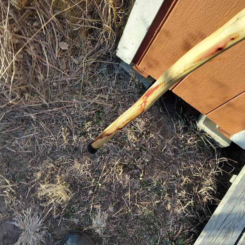 K&ouml;nnte beinhalten: Ein h&ouml;lzerner Wanderstock mit dunkler Gummispitze. Der Stock hat eine hellfarbige Holzmaserung mit rotbraunen Streifen. Der Stock ist &uuml;ber einer Fl&auml;che aus trockenem Gras und in der N&auml;he einer Holzkonstruktion abgewinkelt.