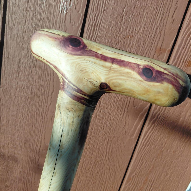 K&ouml;nnte beinhalten: Handgefertigter Holz-Spazierstock mit T-f&ouml;rmigem Griff. Der Griff weist eine helle Holzmaserung mit dunkelvioletten Akzenten und Knoten auf. Der Schaft ist heller. Der Stock ist f&uuml;r Unterst&uuml;tzung und Mobilit&auml;t konzipiert.