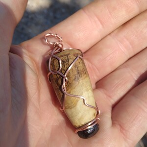 May include: A wooden pendant wrapped in copper wire. The wood is a light brown with darker brown streaks. The pendant is about 2 inches long.