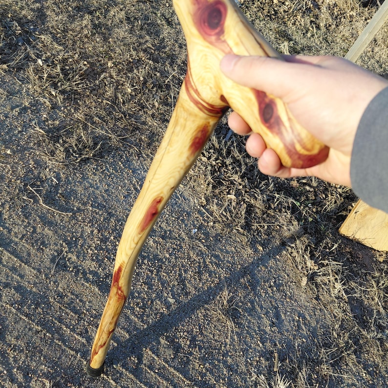 K&ouml;nnte beinhalten: Ein handgefertigter Holzspazierstock mit gebogenem Schaft und rundem Griff. Das Holz hat eine helle, nat&uuml;rliche Farbe mit r&ouml;tlich-braunen Knoten und Akzenten. Der Boden des Stocks hat eine schwarze Gummispitze. Der Stock ist ca. 90 cm lang.