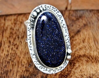 Puede incluir: Un anillo de plata con una piedra preciosa azul engastada en un engaste ovalado texturizado. El anillo tiene una banda texturizada y pequeños detalles plateados.