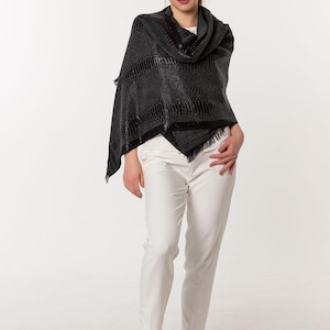 May include: A black patterned shawl with fringing. The shawl is worn over the shoulders. The person is wearing white trousers and beige shoes.