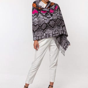May include: A woman wearing a grey, black, and white snakeskin-patterned shawl with orange and pink accents. The shawl has a fringed edge. She is wearing white trousers and black high-heeled sandals.