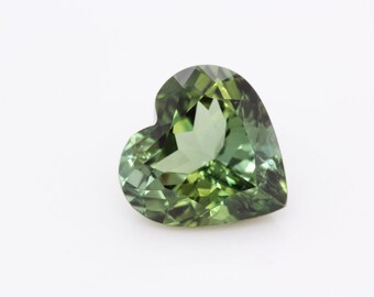 May include: A heart-shaped green gemstone with a faceted cut. The gemstone is set against a white background.