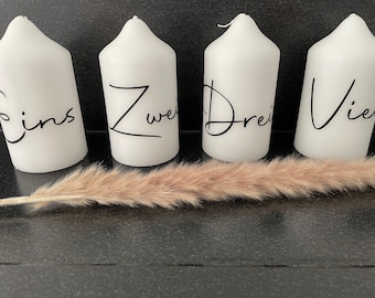 May include: Four white pillar candles with black script, each labeled with a number: Eins, Zwei, Drei, and Vier. A dried, beige ornamental grass stem lies in front of the candles. The candles are on a dark surface.