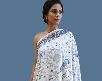 May include: A white shawl featuring intricate grey and blue floral embroidery and peacock motifs, draped over a solid dark blue skirt. The shawl has a wide border with dense botanical patterns and small blue flower accents throughout.