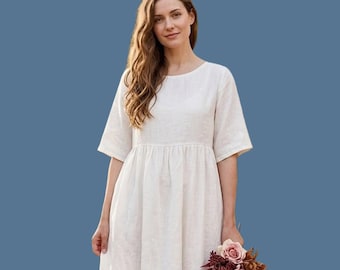 May include: A midi-length dress in an off-white color featuring a round neckline, short sleeves, and a gathered waistline that flows into a loose skirt. The garment is made of a textured fabric and is held alongside a bouquet of dried flowers.
