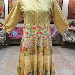 Puede incluir: Un vestido largo amarillo con cuello mandarín y mangas largas. El vestido presenta un estampado floral en rojo, azul y blanco. La falda tiene múltiples niveles con diferentes patrones. El vestido se exhibe en un maniquí.