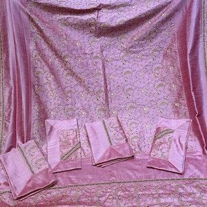 May include: A pink silk bedding set with gold embroidery. The set includes a bedspread and pillowcases, all decorated with floral and paisley patterns. The fabric has a subtle sheen, adding to its luxurious feel.