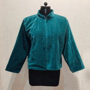 May include: Teal green velvet jacket with a mandarin collar and long sleeves. The jacket is displayed on a black dress form against a neutral background. The jacket has a relaxed fit and a simple, elegant design.