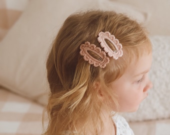 May include: Two pink crocheted hair clips in the shape of an oval, with a scalloped edge. The clips are in a young girl's hair.