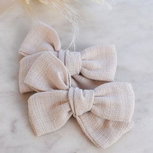 May include: Two beige fabric hair bows tied together. The bows are made of a soft, textured fabric.
